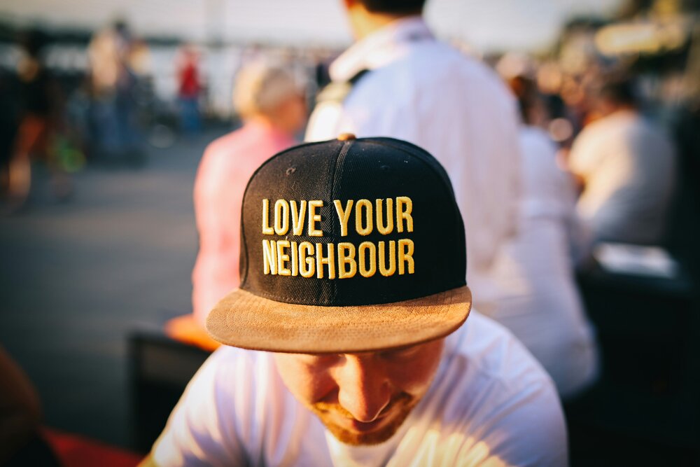 Person wearing a black cap with gold embroidered text reading Love Your Neighbour